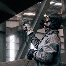 A worker sprays paint on large metal beams in an industrial warehouse.